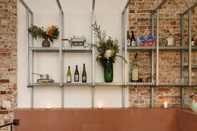 En salle, une structure en acier brut et décorée d'objets de cuisine, bouteilles et bouquets, s'étend au-dessus des tables.