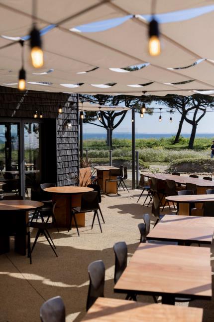 Suspensions lumineuses, assises foncées, tables en bois okoumé et sol sablé pour cette terrasse ouverte sur la nature, que l’on doit au Studio Briand & Berthereau.